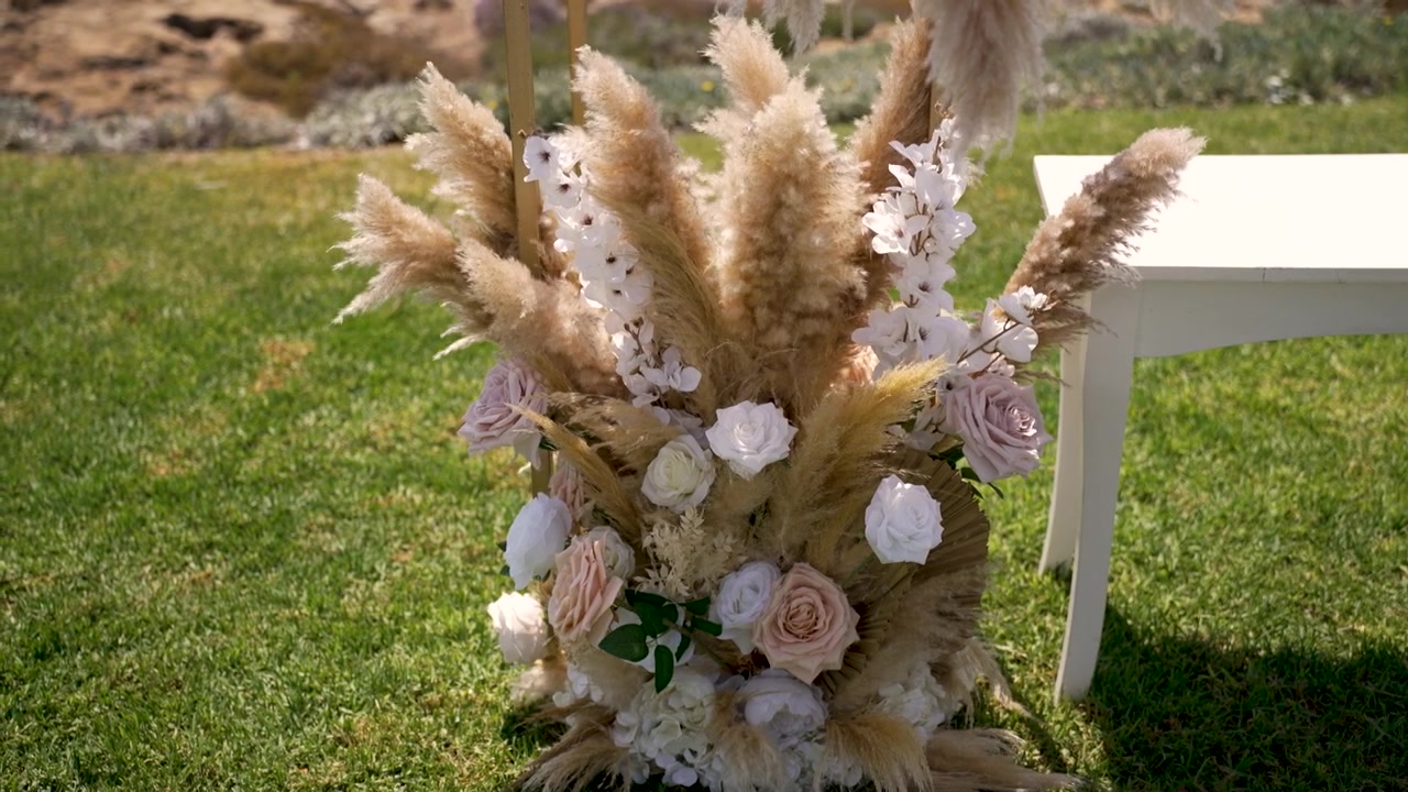 A floral display features pampas grass and roses beside a white table on green grass at an outdoor event. The setting is bright and open with a natural background.