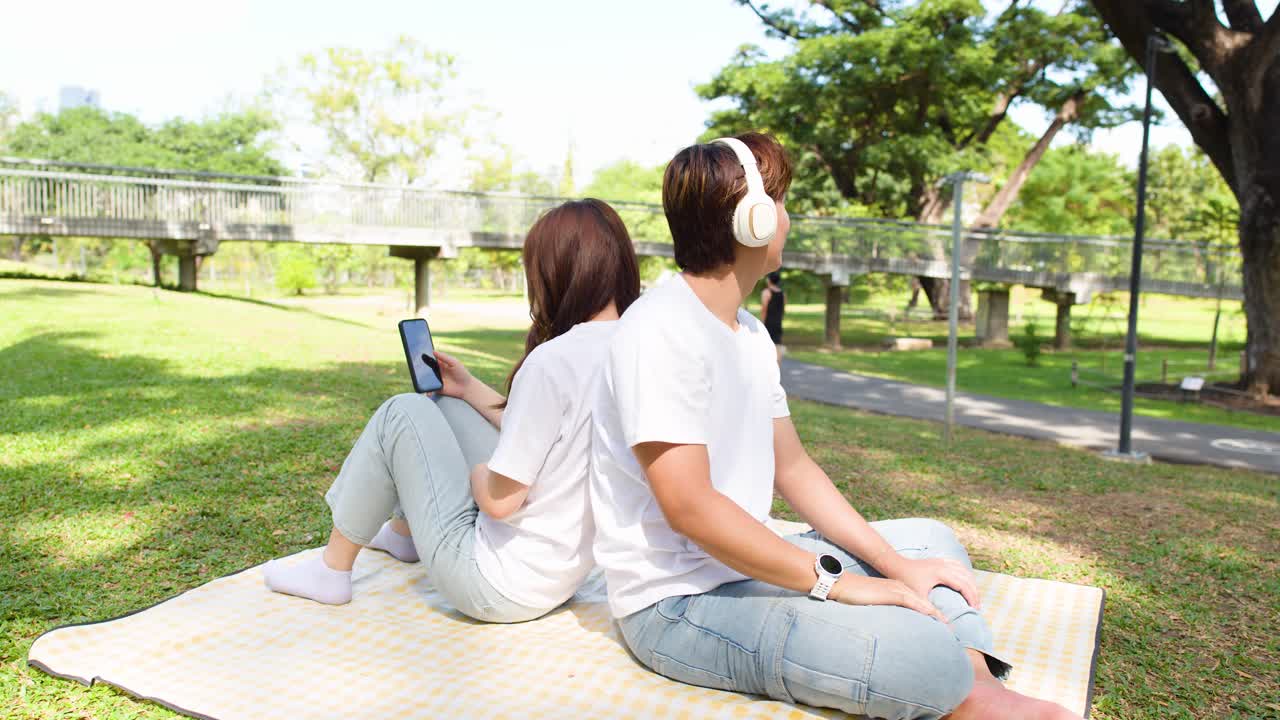 若いカップルがピクニックブランケットの上で背中合わせに座り、ヘッドホンとスマートフォンを公園で共有している。