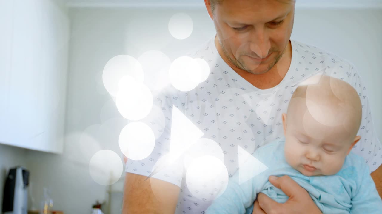 Mutfakta bebek tutan adam, tezgaha uzanıp biberonu kaldırarak besleme yapıyor; bu esnada sağlık temalı bokeh efekti sürükleniyor.
