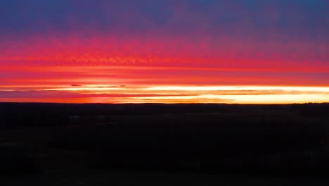 Aerial-view-over-dark-land-while-colorful-golden-hour-sunset-illuminate-sky