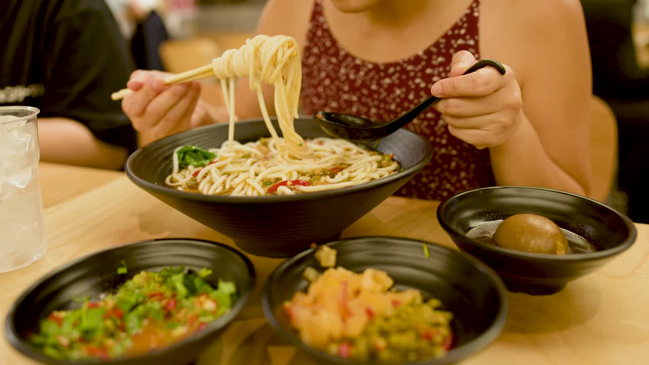 Woman enjoys spicy noodle soup with chopsticks, warm lighting, close-up, casual dining environment
