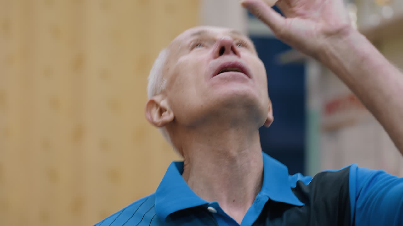 Close up of aged man in blue sportswear concentrating while trying playful trick to keep tennis ball suspended with air breath during indoor practice scene filled with focus and determination