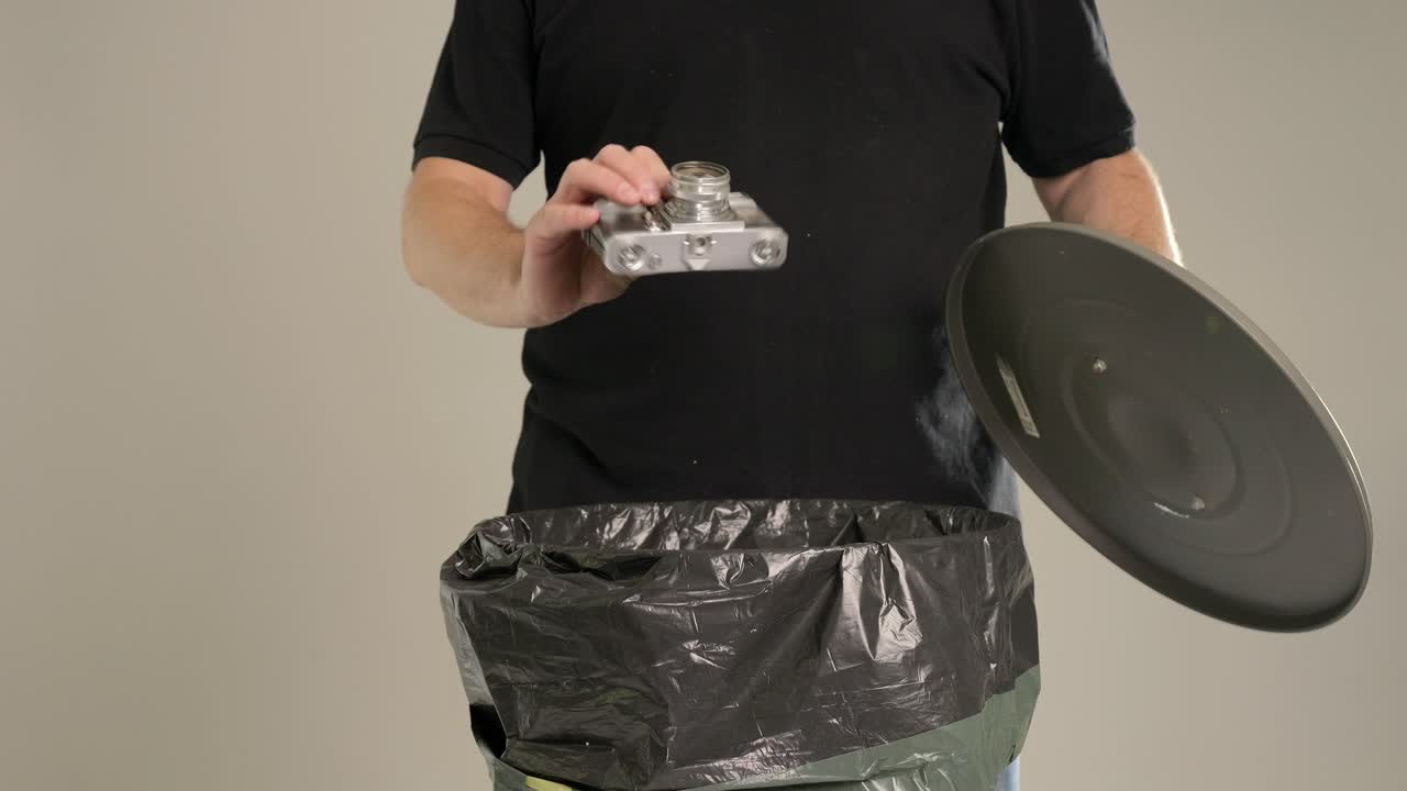 한 남자가 작은, 아주 오래된 금속 카메라를 쓰레기통에 던지고 있습니다.