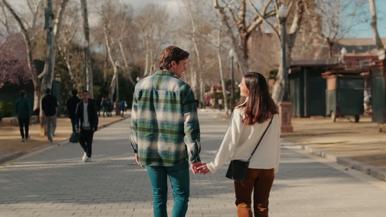 Happy young partners man and woman walking holding hands on the street, back view