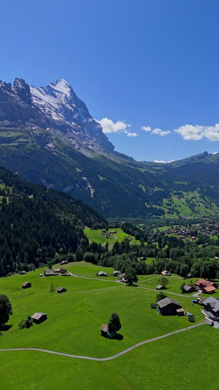Toma cinematográfica vertical de dron haciendo un paneo lento hacia la izquierda desde prados verdes y chalets alpinos para revelar el icónico pico nevado del monte Eiger en Grindelwald, Suiza.