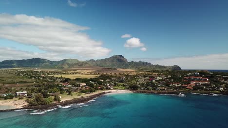 Drone-Alejándose-De-Poipu,-Kauai,-Hawaii,-Playa-Y-Montaña-En-La-Distancia-Sobre-Un-Océano-Azul-Cristalino