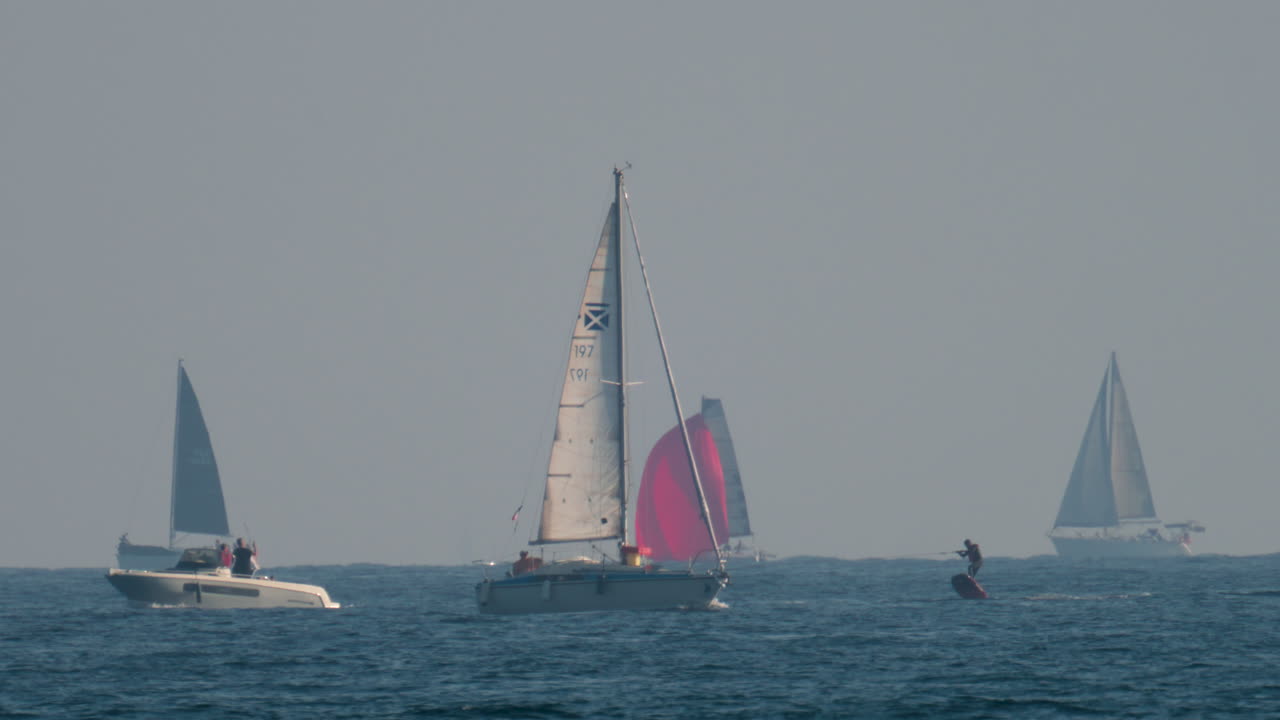 Cannes, France - October 10, 2025: Several sailboats and a catamaran glide over the open sea under a hazy sky, with one striking red sail standing out