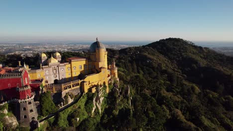 Toma-Aérea-Reveladora-Del-Histórico-Diseño-Arquitectónico-Exterior-Del-Castillo-Del-Palacio-De-Pena-Iluminado-Con-Luz-Solar-En-La-Cima-De-Una-Colina-En-Las-Montañas-De-Sintra-Sobre-La-Ciudad-De-Sintra,-Portugal