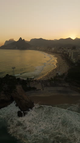 vertical: drone flying over ipanema beach rio de janeiro at sunset, city skyscrapers and mountain silhouettes