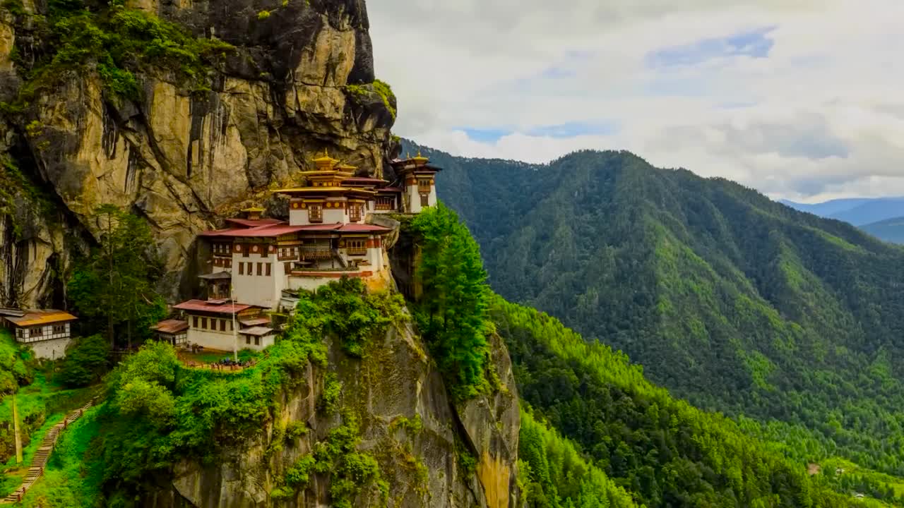 Timelapse footage of the Taktsang Palphug Monastery and the Tiger's Nest on a sharp steep cliff edge during a sunny day while cloud shadows are going over the temple architecture and green nature.
