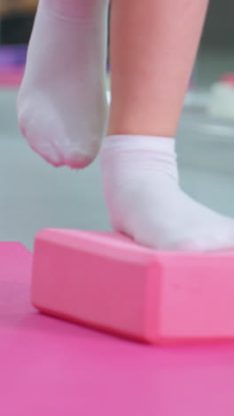 vista de la pierna de una persona con calcetines blancos equilibrándose en un bloque de yoga púrpura durante el entrenamiento de estabilidad en una alfombra de ejercicio rosa, centrándose en la fuerza central, la coordinación y la mejora del equilibrio en un entorno de gimnasio