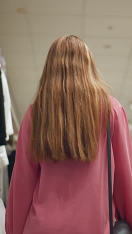vista trasera de la dama llevando una bolsa negra caminando a través de una tienda de ropa mientras toca una tapa negra y se mueve hacia adelante, la tienda está bien iluminada, con ropa bien arreglada en los estantes a ambos lados