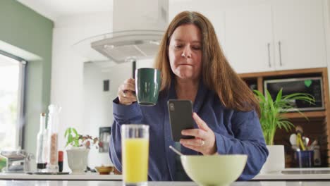 happy caucasian woman eating breakfast, using smartphone in kitchen