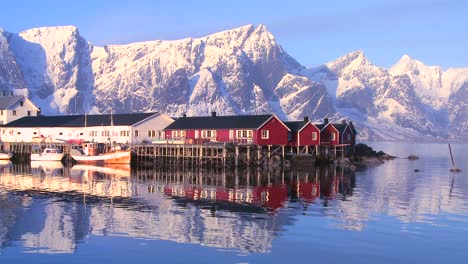 雪地覆蓋的山峰在北極的洛福<unk>群島 (lofoten islands) 的一個紅色漁村後面<unk>起