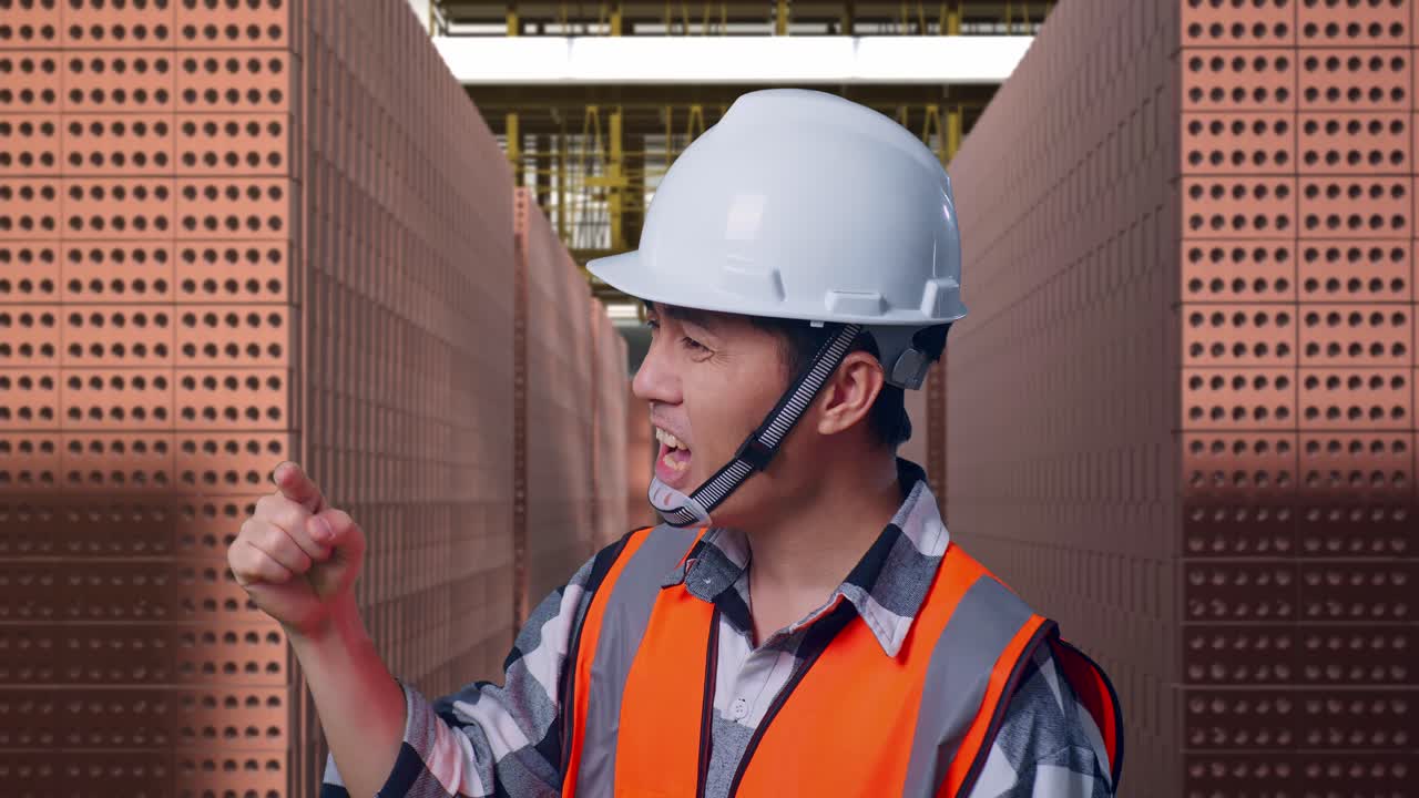 Close Up Side View Of Angry Asian Male Engineer With Safety Helmet Shouting At Someone While Standing With Red Brick Packed in Stacks Are Stored
