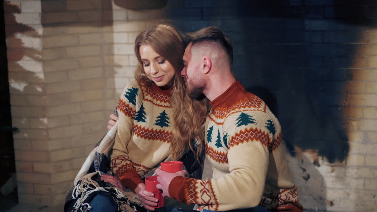 Beautiful loving couple resting near fireplace. Young woman and a man with cups of drink at Christmas. Guy hugging his girlfriend enjoying romantic weekend.