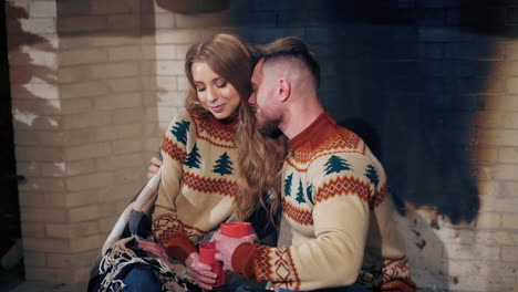 beautiful loving couple resting near fireplace. young woman and a man with cups of drink at christmas. guy hugging his girlfriend enjoying romantic weekend.