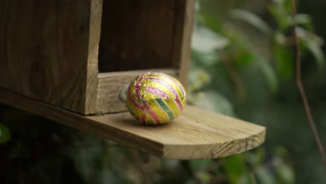 colorful, foil-wrapped chocolate easter egg sits on wooden ledge in natural, outdoor setting, with green foliage in background. captures festive, whimsical charm of easter in a rustic garden location