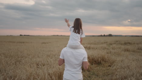 view from the back: daddy carries on his shoulders his beloved little healthy daughter in sun. in slow motion the daughter walks with her father on the field and free and happy waves her hands up.