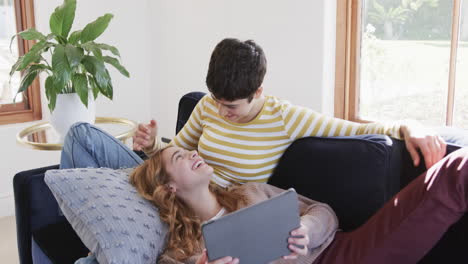 Happy-caucasian-lesbian-couple-lying-on-sofa,-using-tablet-and-laughing-in-sunny-house