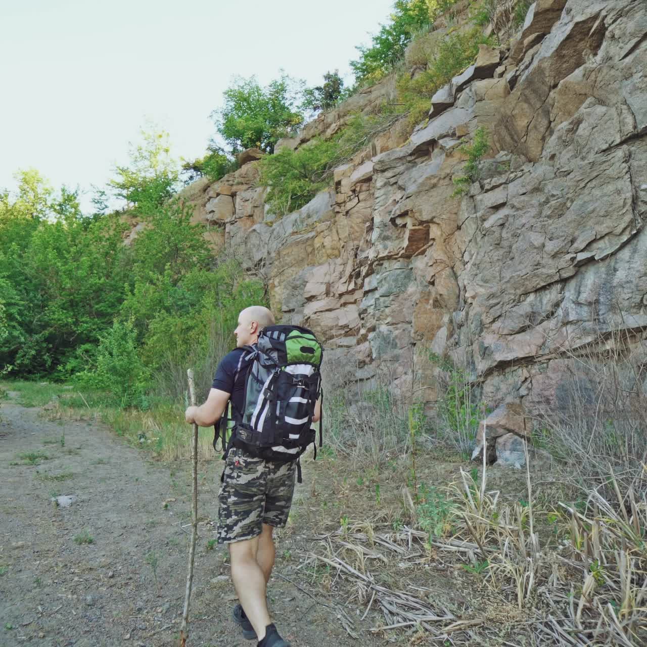 a traveler with back is walking on the path with a stick in his hand near the rocks in the summer day on the background of green trees