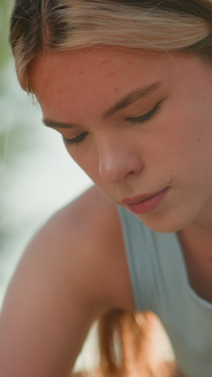 Close-up of young woman slightly bent, focused on something below her, sunlight reflecting off her face, hair tied back, wearing light blue top, with greenery softly blurred in background