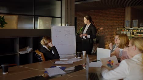 una mujer joven de cabello marrón con gafas redondas y un atuendo profesional está presentando su idea en un puesto durante una reunión de oficina