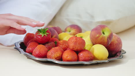 La-Mano-De-Una-Mujer-Elige-Una-Fruta-Deliciosa,-El-Plato-Se-Encuentra-Cerca-De-La-Almohada:-Desayuno-En-La-Cama