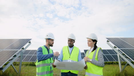 Camera-zooming-on-three-multiethnics-farm-solar-engineers-talking-while-looking-at-blueprint-on-solar-plantation