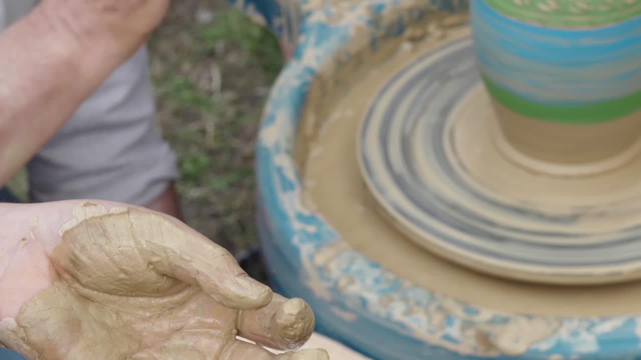 Girl like a traditional potter sits with dirty hands at the potter's wheel. They works outside. Close up hands. Event. Handheld.