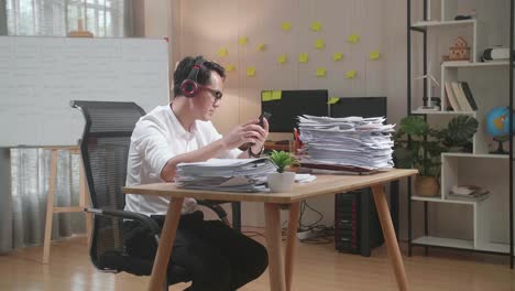 asian man with headphones listening to music on smartphone after working with documents at the office