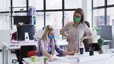 Diverse-female-office-colleagues-wearing-face-masks-discussing-over-blueprint-plan-at-modern-office