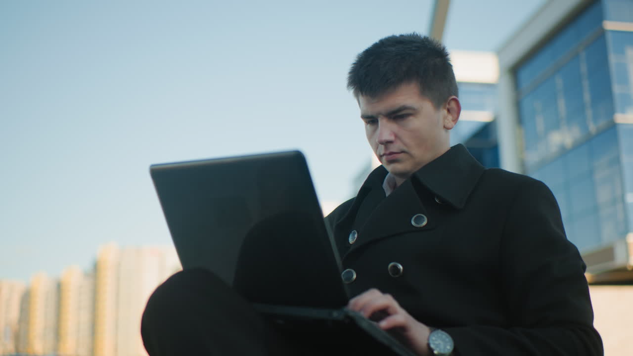 흑색 코트를 입은 부동산 거장, 도시 환경에서 노트북을 운영하는 데 집중, 손목에 보이는 손목시계, 현대적인 사무실 건물과 배경의 도시 요소