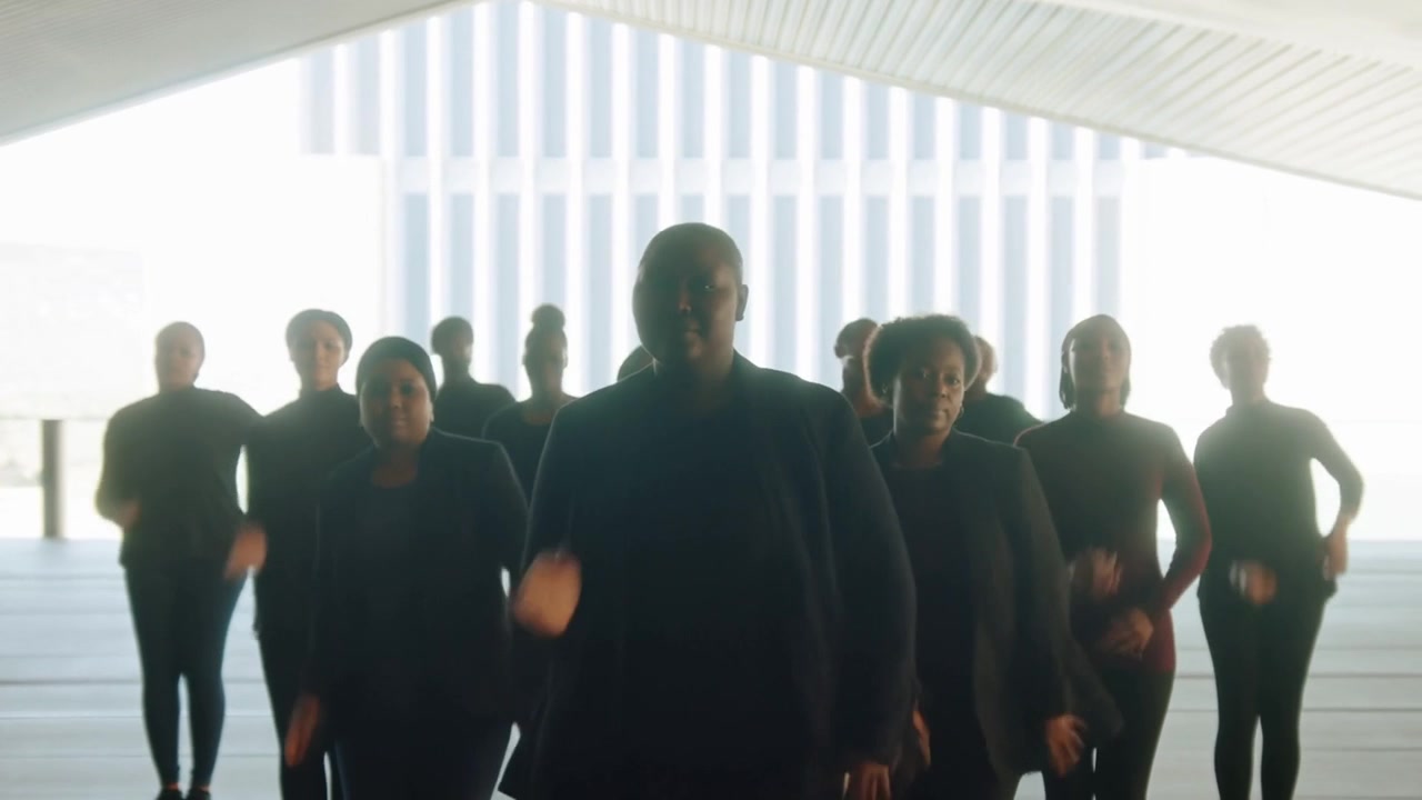 Un leader centrale che guida avanza con il braccio che oscilla sotto un baldacchino, dando il segnale a un ensemble che indossa blazer.