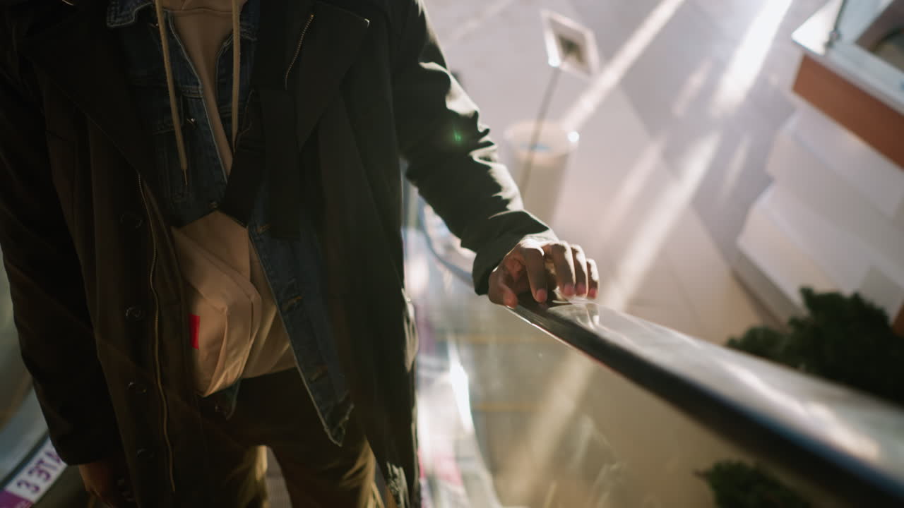 Close-up of young man with hoodie, hand taping on escalator railing, moving through modern shopping mall with soft lighting and bright surroundings, small bag over shoulder