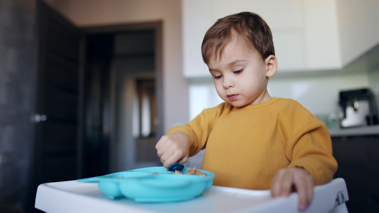 Lovely toddler in orange sweater eating porridge. Baby boy chewing food and waving his spoon in the air.