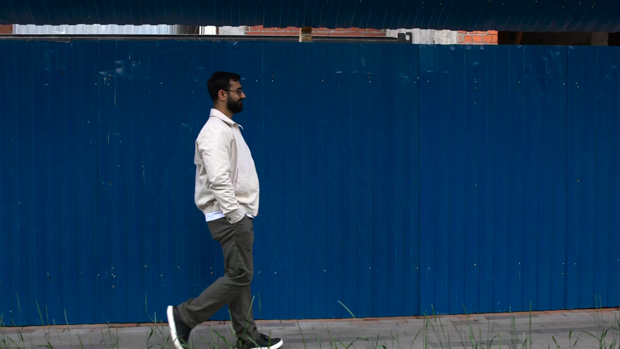 Yan profilden yetişkin bir adam, asfalt kaldırım boyunca, yıpranmış metal dokulu, büyük mavi oluklu bir çitin yanından geçiyor. Üstte tuğla yapılar, altta seyrek çim, serin gün ışığı ve sessiz, kapalı bir ortam.