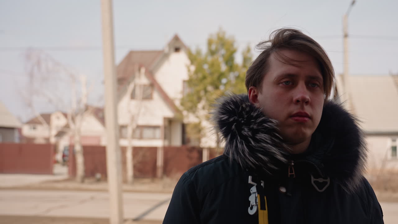 Two young men walks outdoors in residential neighborhood, one wearing winter jacket with fur hood looking ahead seriously, other wearing glasses and backpack partially visible, urban casual lifestyle