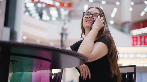 hermosa mujer charlando por teléfono con camiseta negra y gafas, mirando hacia arriba con un fondo borroso con efectos de luz bokeh y estructuras modernas