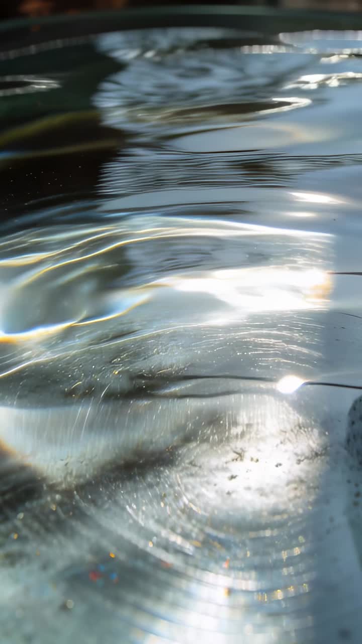 Vertical video: Droplet hitting shallow water surface at basin, creating ripples and sun caustics