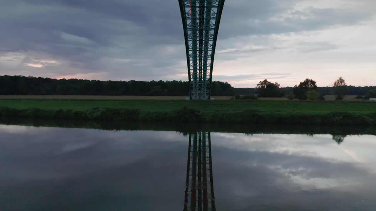 Flying under gas pipes across calm river Morava. Aerial sunset footage of a gas pipe bridge reflecting in water.