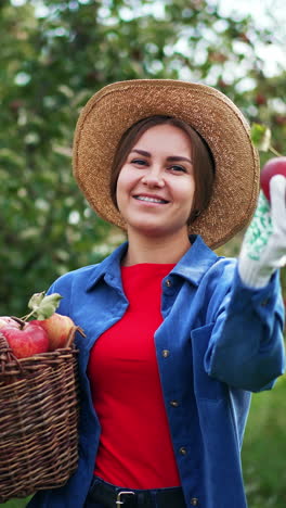 happy female farmer holding a basket of ripe apples. smiling lady offering a red fruit to the camera. garden in blur at backdrop. vertical video