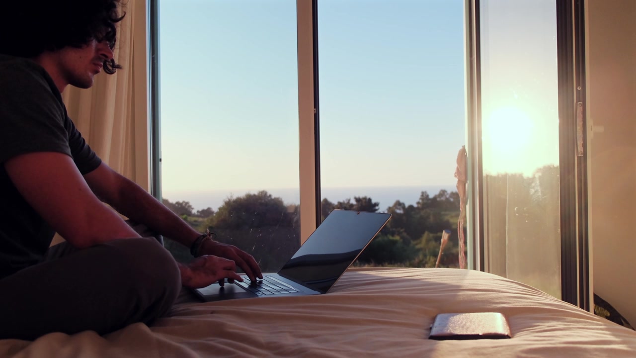 Digital nomad on bed with laptop, enjoying ocean and beach view
