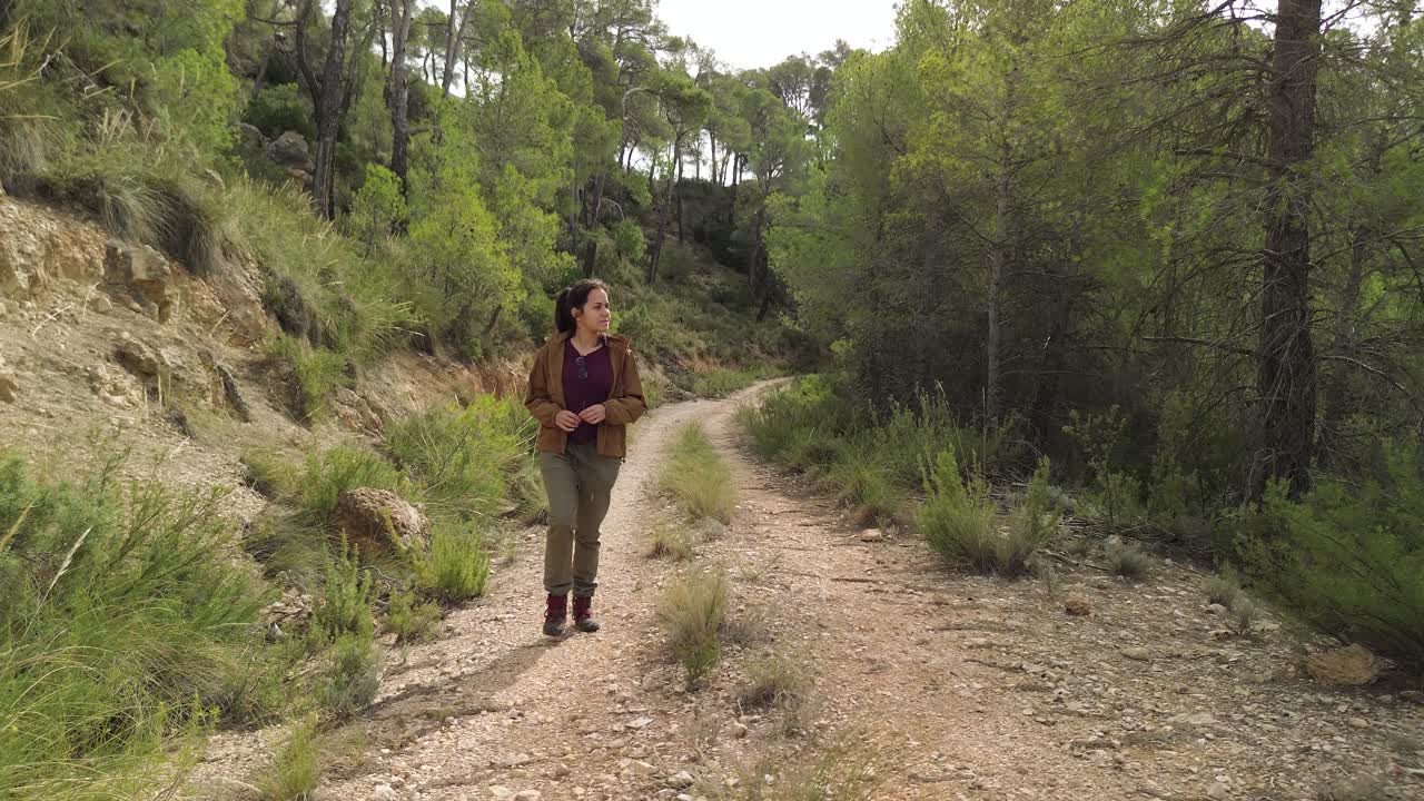 Ripresa frontale con drone di un'escursionista bruna rilassata che cammina lungo un sentiero nel bosco circondata dalla vegetazione