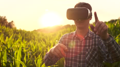 A-male-farmer-in-virtual-reality-glasses-wearing-a-plaid-shirt-and-jeans-in-a-field-at-sunset-controls-the-corn-irrigation-system.-Modern-farmer-new-technology-farmer-of-the-future