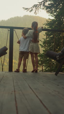 brother and sister watch nature of calm morning forest hugging on balcony of glamping house. boy and girl together rest enjoying deep atmosphere