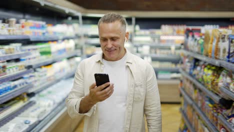 A-middle-aged-man-walks-through-a-grocery-store-talking-on-a-video-link.-Headphones-in-the-ears.-Shopping-trip.-Modern