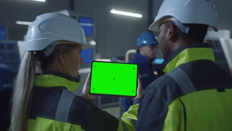 industry 4.0 factory: chief engineer and project supervisor in safety vests and hard hats, talk, use digital tablet computer with green screen, chroma key. workshop with machinery. zoom out