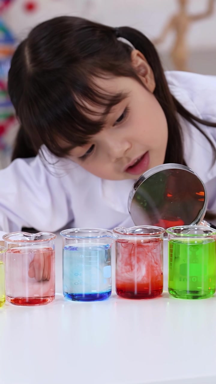 Gadis muda menggunakan kaca pembesar untuk mengamati cairan berwarna-warni di dalam gelas kimia selama pelajaran sains di kelas