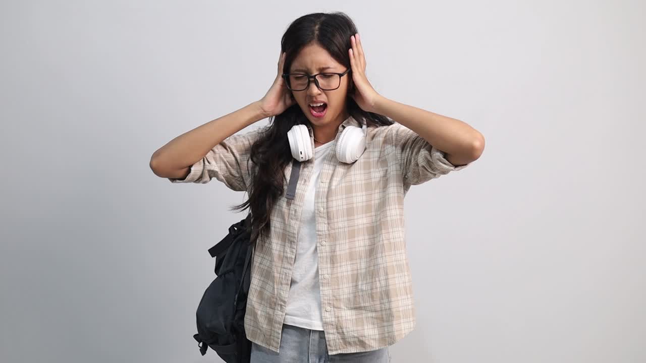 Una giovane studentessa universitaria asiatica infelice, triste e arrabbiata chiude gli occhi e si copre le orecchie con le mani, infastidita dal rumore forte, su sfondo bianco isolato da studio, emozioni, avversione ai suoni forti.